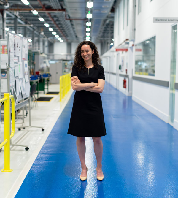Une ingénieure dans une usine de fabrication mécatronique, avec des lignes de production automatisées de haute technologie à l'arrière-plan.