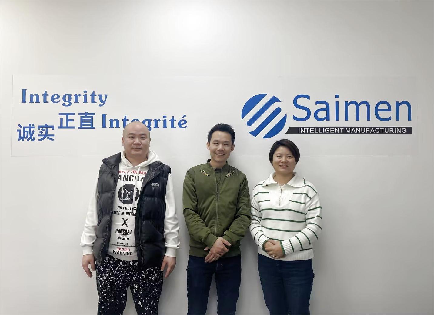 Saimen team with visiting clients in front of a wall showcasing the company’s commitment to integrity, highlighting successful global partnerships.