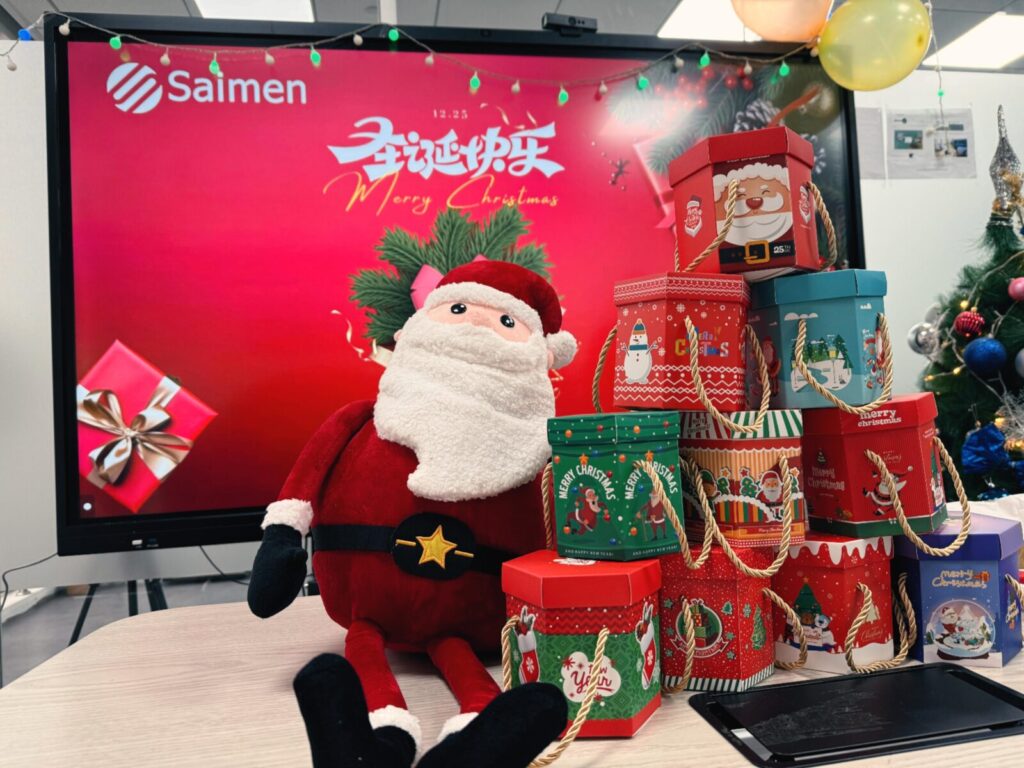 A festive office setting with a large monitor displaying 'Merry Christmas' in Chinese, surrounded by colorful Christmas gift boxes and a plush Santa Claus.