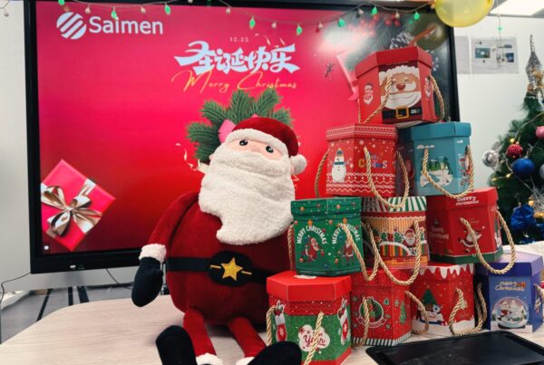 A festive office setting with a large monitor displaying 'Merry Christmas' in Chinese, surrounded by colorful Christmas gift boxes and a plush Santa Claus.